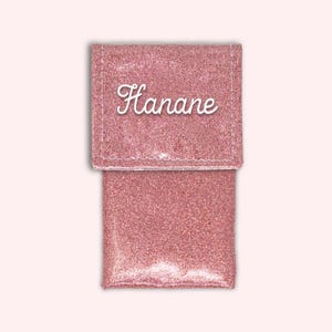 Puede incluir: Una bolsa de cosm&eacute;ticos de color rosa con purpurina y una solapa frontal transparente. La bolsa tiene el nombre "Hanane" escrito en blanco en la parte delantera.