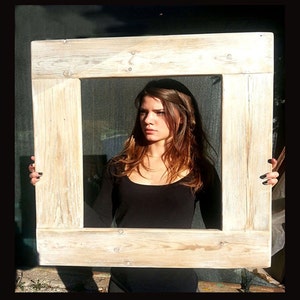 Op de afbeelding: Een vrouw houdt een groot, witgeschilderd houten frame voor haar gezicht. Het frame is gemaakt van gerecycled hout en heeft een rustieke, verweerde uitstraling.