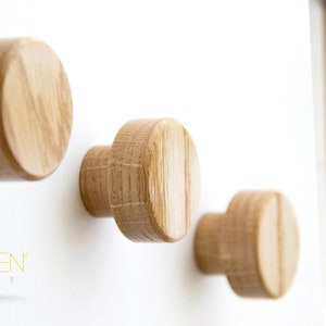 May include: Three wooden wall hooks in a circular shape. The hooks are made of natural wood and have a light brown color. The hooks are mounted on a white wall.