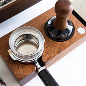 Puede incluir: Un soporte de madera para tamper de espresso con un portafiltro de acero inoxidable y un tamper negro y marrón. El tamper tiene el texto "Wooden Art" en él.
