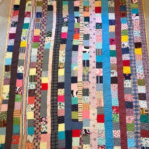 May include: A rectangular quilt constructed from vertical strips of multi-colored fabric squares. The quilt features a diverse array of patterns, including floral, plaid, striped, and solid designs in various colors, finished with a yellow border.