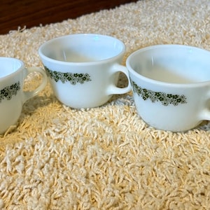 May include: Five white teacups with a green floral pattern around the rim. Each cup has a handle and is set on a cream-colored, textured surface. The cups are arranged in a slightly staggered row.