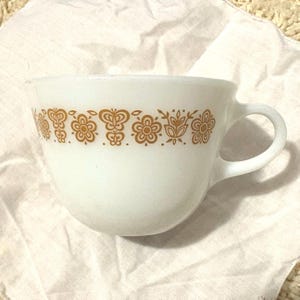 May include: White glass measuring cup with a handle and a decorative band of gold-colored floral and butterfly designs. The cup has a rounded shape and a pouring spout.