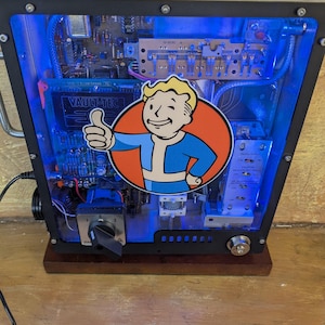 May include: A custom-built electronic device with a clear front panel, showcasing internal components illuminated by blue LED lights. A Vault-Tec logo sticker is prominently displayed. The device has a black frame and sits on a wooden base.