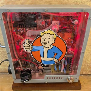 May include: A gray and clear electronic device with red internal lighting. The device features a Vault-Tec logo sticker with a thumbs-up character. The text "VAULT-TEC INDUSTRIES" is visible on the device.