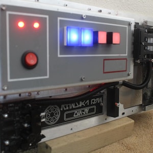 May include: A custom-built electronic panel with a gray and silver finish. It features red and blue lights, a red button, and a black panel with text and a symbol. The panel is mounted on a wooden base.