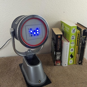 May include: A silver-colored, retro-style lamp with a circular face featuring blue lights and a red and silver trim. The lamp is on a black base, with a row of books to the right.