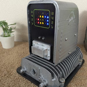 May include: A silver-colored, retro-style device with a black front panel featuring a grid of colorful lights and the word "Firefly". The device has a rectangular base and a ribbed, industrial design.