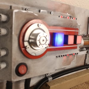 May include: A detailed, weathered metal panel with red and silver accents, featuring a circular design and a glowing blue cube. The panel includes a red power button and a black, corrugated tube. The text "ANIO" is visible.