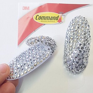 May include: Two silver Command brand hooks covered in small, sparkling rhinestones. One hook is curved, and the other is oval-shaped. The hooks are on a white background with the Command brand packaging visible.