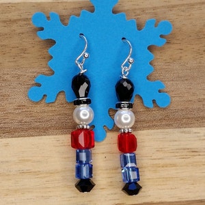 May include: A pair of dangle earrings featuring a beaded design that resembles a toy soldier. The earrings are made with black, white, red, and blue beads. The earrings have silver hooks.