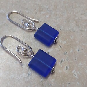 May include: A pair of earrings with square, cobalt blue glass beads. The earrings have silver-colored, curved ear wires and small decorative beads at the base of the blue squares. The earrings are displayed on a light-colored, textured surface.