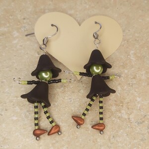 May include: A pair of witch earrings made with black and green beads. The witches have black hats, green faces, and black and green striped legs. The earrings are attached to silver hooks.