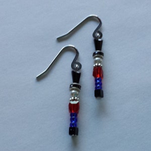 May include: A pair of dangle earrings featuring a soldier design. Each earring has a silver hook and a beaded soldier figure with a black hat, red and blue accents, and white beads. The earrings are on a white background.