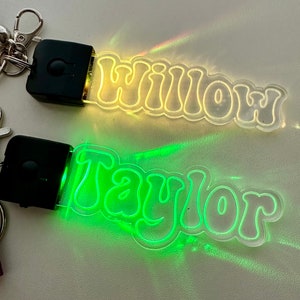 May include: Two personalized keychains with illuminated names. One keychain glows yellow with the name "Willow", the other glows green with the name "Taylor". Each keychain has a black rectangular light source and a silver clasp.