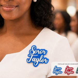May include: A blue name badge with white script lettering reading Soror Taylor, pinned to a white garment. A small rectangular graphic in the corner displays three additional name badge color combinations in blue, red, and green with white text.