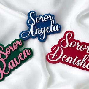 Puede incluir: Tres etiquetas de nombre personalizadas con la palabra "Soror" seguida de un nombre. Las etiquetas son de acrílico y son de color verde, azul y rojo.