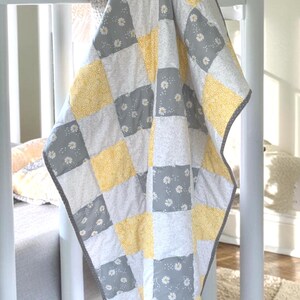 May include: A patchwork quilt featuring a repeating grid of square fabric panels. The design alternates between solid yellow floral patterns, grey squares with white daisy prints, and light grey textured squares, all finished with a grey border.