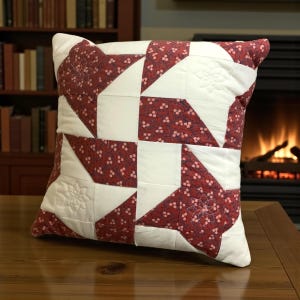May include: Square decorative pillow with a patchwork design. The pillow features a pattern of red floral fabric and white fabric, with embroidered floral details. The pillow is placed on a wooden table.