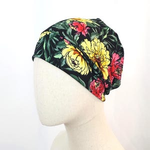Könnte beinhalten: Eine schwarze Beanie-Mütze mit einem lebendigen Blumenmuster mit gelben, roten und rosa Blumen mit grünen Blättern. Die Mütze ist aus einem weichen, flexiblen Material gefertigt und passt sich der Kopfform an.