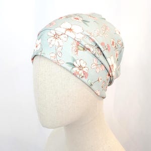 Può includere: Un berretto blu chiaro con un motivo floreale. Il cappello presenta un motivo ripetuto di fiori bianchi e rosa con foglie verdi. Il cappello è realizzato in un tessuto morbido ed elastico ed è mostrato su una testa di manichino.
