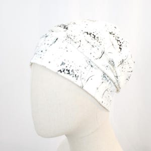 May include: A white beanie hat with a black and gray floral pattern. The hat is made of a soft material and is designed to fit snugly on the head. The pattern includes small, scattered dots and delicate, curved lines.