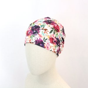 May include: A white floral print beanie with a gathered design. The beanie features a variety of pink, purple, and orange flowers with green leaves.