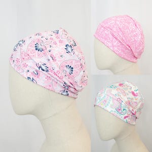 Può includere: Tre copricapi fantasia rosa e bianchi sono esposti su teste di manichini. I motivi includono disegni floreali, geometrici e paisley. I copricapi sono probabilmente per scopi medici o di moda.