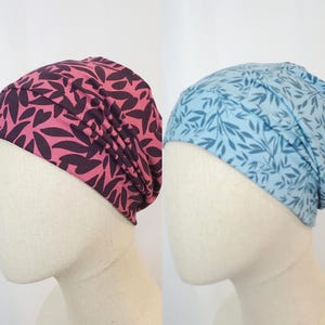 Può includere: Due copricapi in tessuto sono esposti su teste di manichino. Uno è rosa con forme di foglie viola scuro, e l'altro è azzurro con un motivo a foglie blu più scuro. I copricapi sono raccolti nella parte superiore.