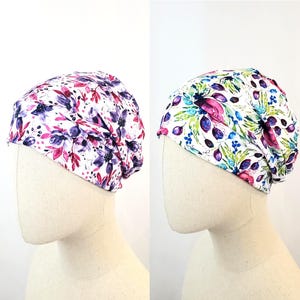 May include: Two floral print beanies on mannequin heads. The beanie on the left has a white background with purple and pink floral patterns. The beanie on the right has a white background with blue, green, and pink floral patterns.