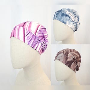 Può includere: Tre copricapi colorati sono esposti su teste di manichini. Uno è decorato con strisce verticali rosa e viola. Un altro è un design blu tie-dye. Il terzo presenta un motivo geometrico nei toni del marrone.