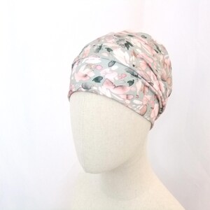 Puede incluir: Un cubrecabezas con estampado floral en tonos grises, rosas y verdes. La tela es suave y drapeada, con un diseño tipo turbante. El cubrecabezas se muestra sobre una cabeza de maniquí sobre un fondo blanco.