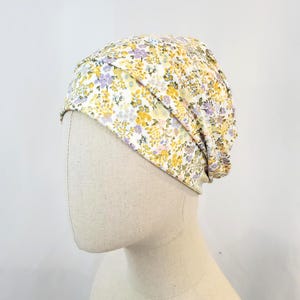 May include: A floral print beanie hat in yellow, purple, and white. The hat is made of a soft fabric and is displayed on a mannequin head. The beanie features a delicate pattern of small flowers.