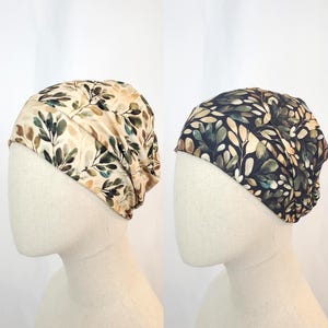 Puede incluir: Dos gorros de tela con estampados de hojas. Un gorro tiene un fondo beige con hojas verdes y marrones. El otro gorro tiene un fondo azul marino con hojas verdes y beige. Ambos gorros se muestran en cabezas de maniquí.