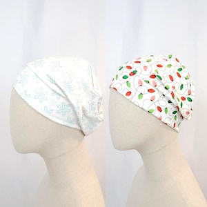 Può includere: Due berretti in tessuto su teste di manichini. Un berretto è bianco con fiocchi di neve azzurri. L'altro berretto è bianco con un motivo di luci natalizie rosse e verdi. Entrambi i berretti sono realizzati in tessuto morbido ed elastico.