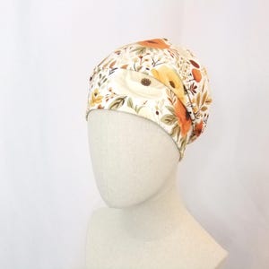 Könnte beinhalten: Eine weiße Beanie mit floralem Muster mit orangefarbenen und gelben Blumen. Die Beanie ist aus weichem, dehnbarem Stoff gefertigt und perfekt, um Ihren Kopf warm und stylisch zu halten.