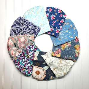 Può includere: Una disposizione circolare di dieci mascherine in tessuto con vari motivi, tra cui blu scuro con foglie bianche, azzurro con piccoli fiori, blu scuro con fiori rosa, blu con stampe botaniche e diversi disegni floreali.