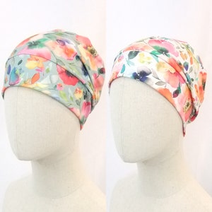 Pode incluir: Dois gorros de malha com estampa floral, um com fundo verde claro e o outro com fundo branco. Ambos os gorros apresentam uma estampa floral em estilo aquarela com flores rosa, laranja e amarelas.