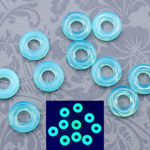 May include: A selection of light blue, ring-shaped beads. The beads are translucent with a central hole. A smaller image shows the beads glowing green under a black light. These beads are suitable for jewellery making.