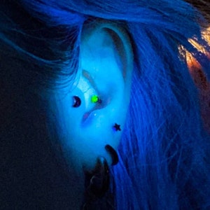 May include: A close-up of a person's ear with multiple piercings. The ear is pierced with a green gem, a black star, and a black crescent moon. The person has blue hair.