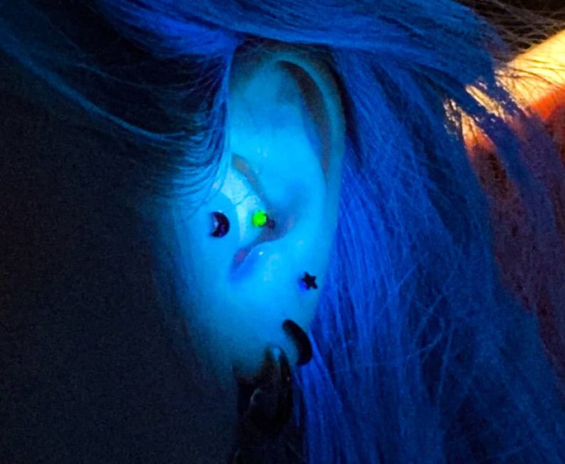 May include: A close-up of a person's ear with multiple piercings. The ear is pierced with a green gem, a black star, and a black crescent moon. The person has blue hair.