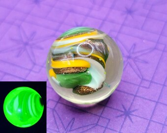 May include: A clear glass paperweight with colorful swirls of green, yellow, brown, and white. The paperweight has a glittery, textured appearance. A small, glowing green orb is in the lower left corner. The background is purple with a star pattern.