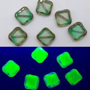 May include: A collection of seven square-shaped beads. The beads have a light green hue with a metallic bronze-colored edge. The bottom half of the image shows the same beads glowing bright green against a dark blue background.