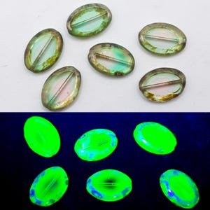 May include: A collection of oval-shaped glass beads with a silver-colored rim. The beads have a translucent center with hints of pink and green. The bottom half of the image shows the same beads glowing bright green under a black light.
