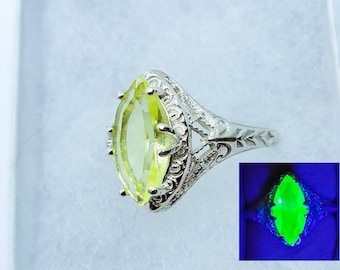 May include: A silver ring with a yellow marquise-shaped gemstone. The ring has a delicate filigree design. The gemstone glows green under a black light.