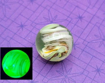 May include: A clear glass marble with a swirling design of white, gold, and yellow. The marble is set against a purple background with a star pattern. A smaller, glowing green marble is in the corner.