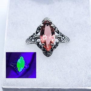 May include: A silver ring with a pink marquise-shaped gemstone set in a filigree design. The gemstone glows green under a black light.