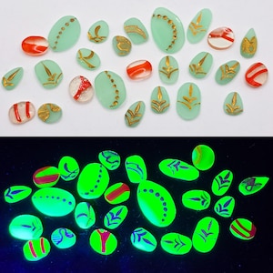 Può includere: Una collezione di piccoli cabochon in vetro lisci e ovali in verde menta pallido, trasparente e arancione. Molti presentano motivi dipinti in oro metallizzato, inclusi punti e disegni a foglia. Sotto la luce UV, il vetro brilla di verde neon.