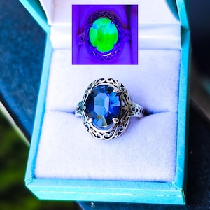May include: A silver ring featuring an oval-cut dark blue gemstone set in an intricate filigree metal band. An inset shows the same ring under UV light, where the gemstone glows with a bright neon green color, displayed in a light blue box.