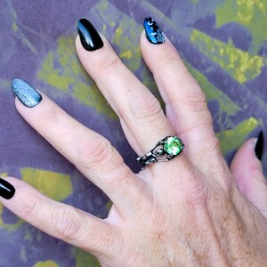 May include: A hand wearing a silver ring with a green gemstone. The ring has a skeletal design. The hand is also wearing black and silver nail polish.